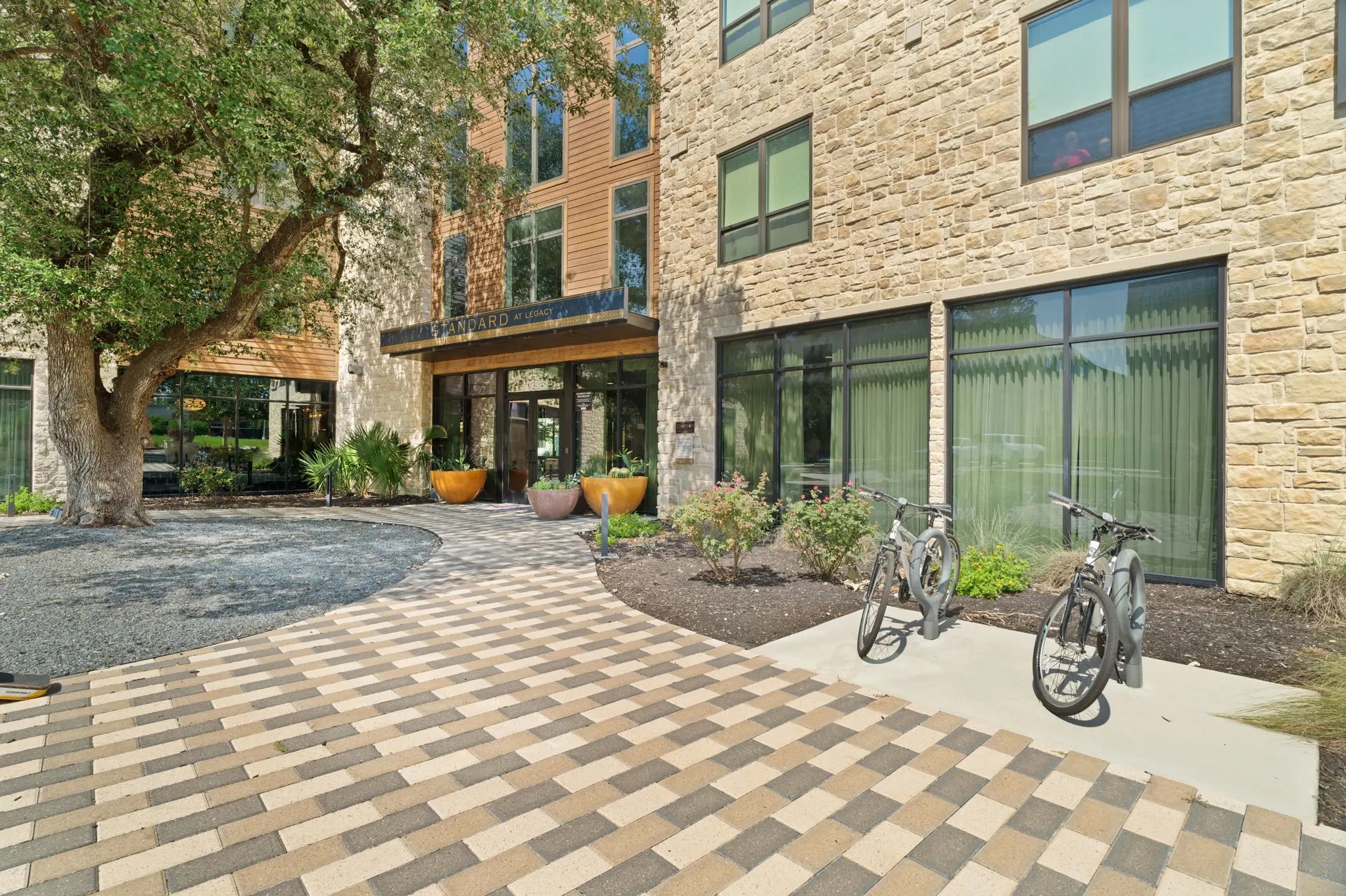 Sunlit stone building courtyard with patterned pavers, trees, planters, large windows, and two bicycles; a welcoming, modern outdoor space.
